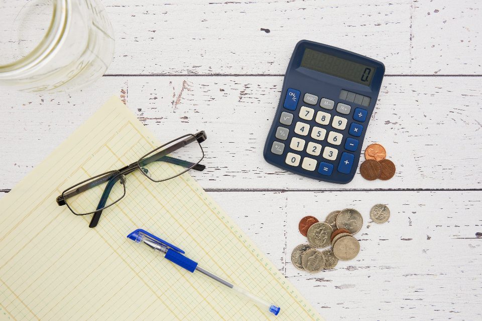 A calculator, glasses, coins and a notepad on a white wooden desk